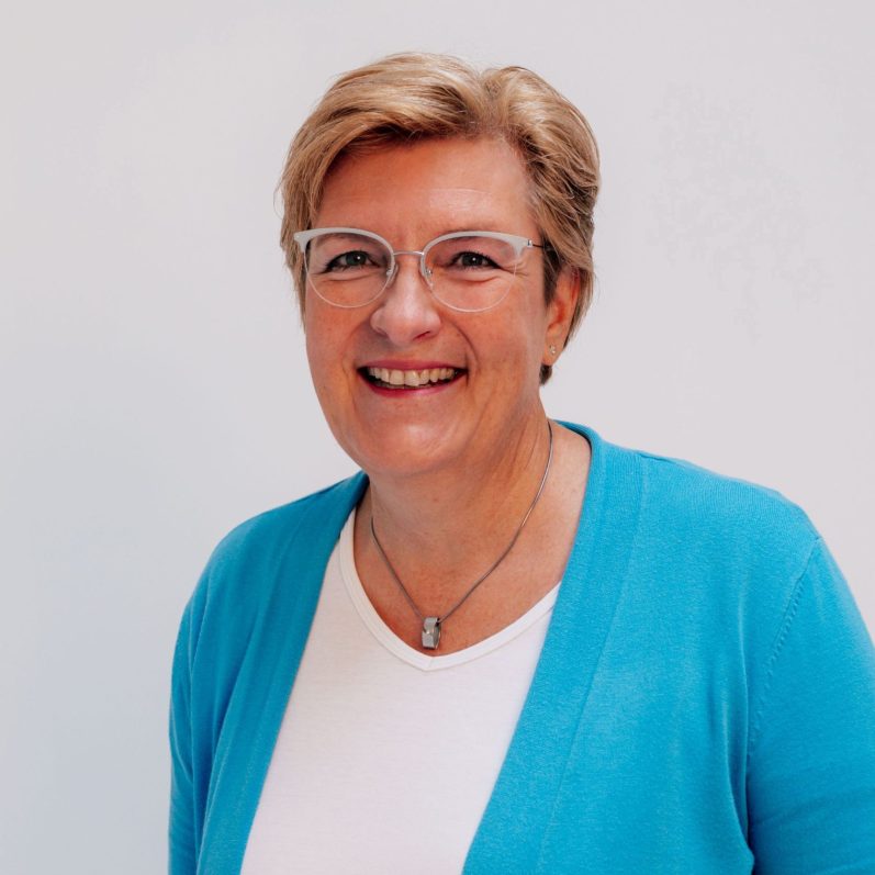 Portrait Jutta Krämer Frau mit blonden Haaren, Brille und blauer Jacke, lächelt freundlich.